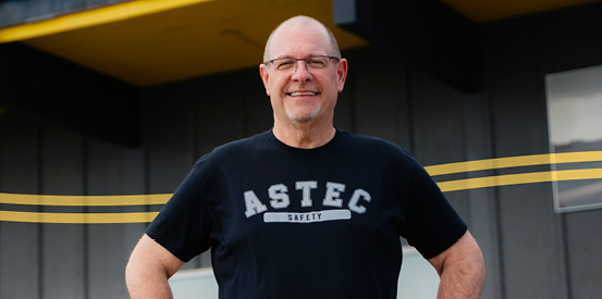 A person wearing a black ‘ASTEC SAFETY’ t-shirt stands in front of a building with yellow lines on the ground.