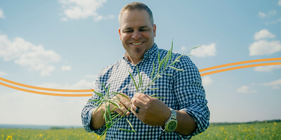 An individual wearing a checkered shirt, stands in a field holding green plant stalks. Two orange lines curve behind their midsection.