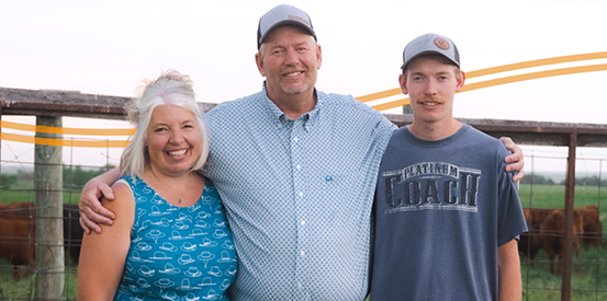 A group of three people standing together, smiling in front of a cattle fence with some cows visible behind them. An orange wavy line runs behind the group.