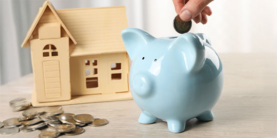 House model and money on wooden table. Person putting coin into piggy bank indoors.